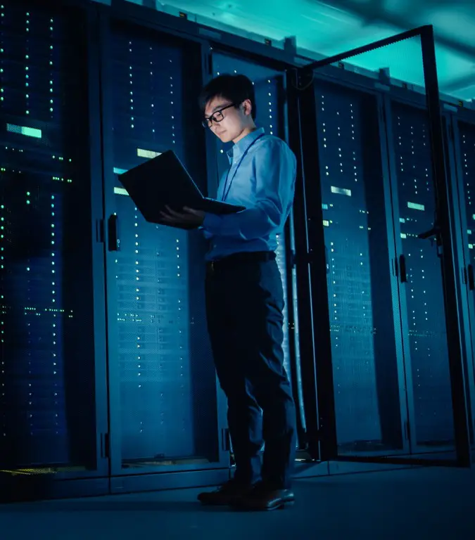 Man standing while looking at a laptop in a room of servers